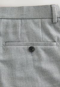 Pantaloni in tessuto grigio con trama texturizzata, una piccola tasca posteriore con bottone nero e pieghe discrete vicino alla cintura.