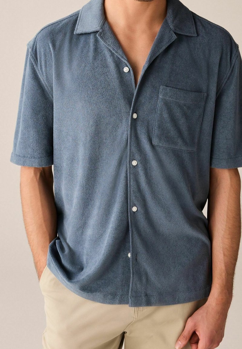 Lyhythihainen nappitettava paita pehmeää, sinistä pyyhekuosista kangasta, jossa on teräväkaulus, tasku ja valkoiset napit. Rentoutunut istuvuus ja teksturoitu pinta.