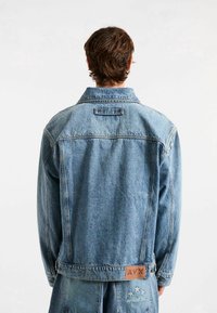 Hellblaues Denim-Jacket mit einem standardisierten Kragen, Knopfverschluss und einem gebrandeten Lederpatch auf dem Rücken. Abgenutzte Textur und lässige Passform.