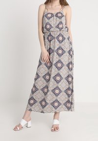 Maxi šaty s geometrickým vzorem v námořnické modré a krémové barvě, tenké ramínka, nařasený pas a splývavá sukně. Oblečené s bílými blokovými podpatky.