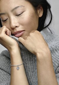 Bracciale d'argento con un charm a forma di cuore con accenti blu, indossato su una mano contro un maglione grigio testurizzato. Inquadratura ravvicinata.