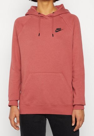 Persoon draagt een stofroze Nike hoodie met voorzak en zwart logo, gecombineerd met een zwarte broek, tegen een effen lichte achtergrond.