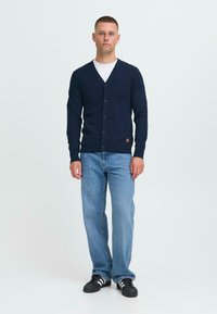 Férfi áll egy tengerészkék kardigánban, fehér inggel, kék farmernadrággal és fekete Adidas tornacipővel, fehér csíkokkal, egy sima fehér háttér előtt.