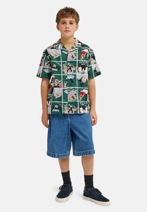 Junge steht, trägt grünes kurzärmeliges Hemd mit Comic-Panels, blaue Jeansshorts, schwarze Socken und dunkle Sneakers vor weißem Hintergrund.