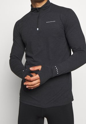 Mann trägt ein eng anliegendes dunkelgraues langärmliges Sportoberteil mit Daumenlöchern und einem Viertel-Reißverschluss, steht mit verschränkten Händen.
