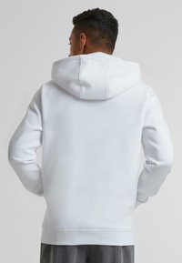 Hvid pullover-hoodie med en rummelig hætte, ribbede manchetter og talje; lavet af blødt stof, glat tekstur og minimale sømme på ryggen.