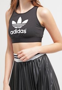 Top cropped nero realizzato in materiale elasticizzato con il logo Adidas bianco; abbinato a una gonna nera a pieghe con una vita a righe.