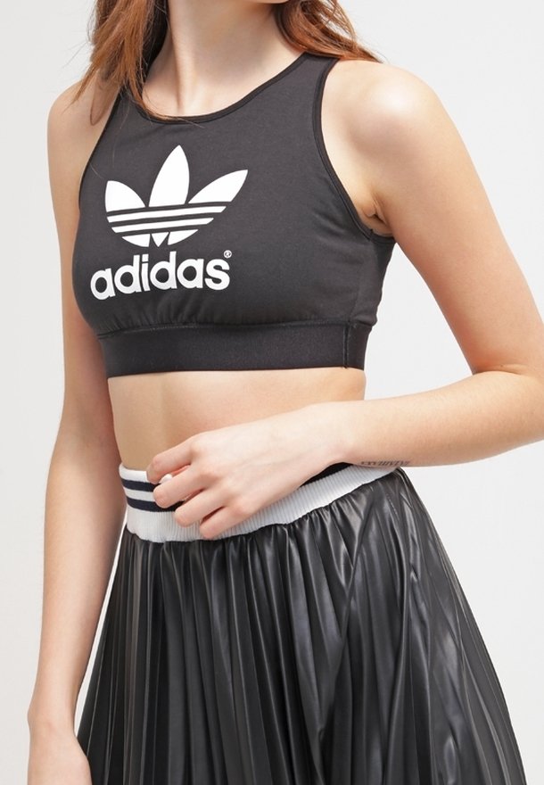 Top cropped nero realizzato in materiale elasticizzato con il logo Adidas bianco; abbinato a una gonna nera a pieghe con una vita a righe.