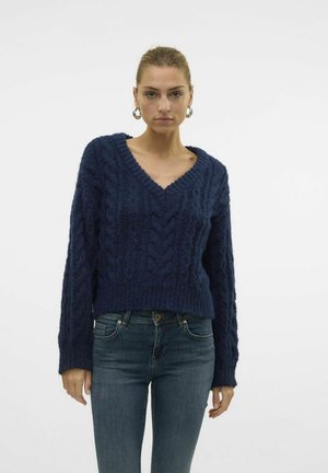 Pull en tricot bleu marine avec un col en V, motif en relief en câble et manches longues, associé à un jean bleu foncé.