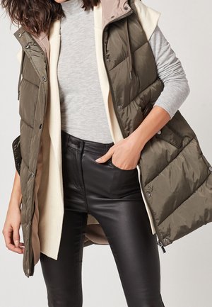 Femme portant un col roulé gris, un cardigan crème, un gilet matelassé vert olive et un pantalon en cuir noir, main dans la poche, sur un fond uni.