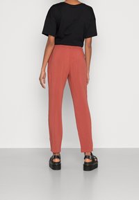 Persoon in losse roestkleurige broek, zwart t-shirt met korte mouwen en zwarte plateausandalen, staand tegen een witte achtergrond.