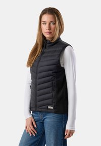 Mladá žena s dlhými svetlohnedými vlasmi, oblečená v čiernej prešívanej bezrukávovej veste cez bielu dlhú blúzku a modré džínsy, stojaca pred jednoduchým pozadím.