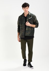 Jasje met camouflagedessin in donkergroen, lichtgewicht materiaal met knoopsluiting, gecombineerd met olijfgroene broek en zwarte sneakers met witte zolen.