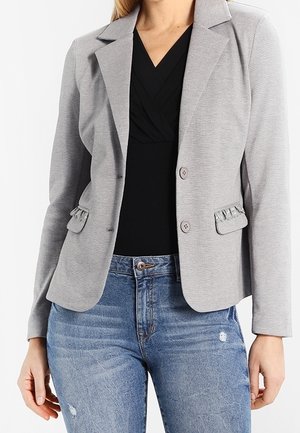 Femme portant un blazer gris clair ajusté à deux boutons avec des poches à rabat froncées, sur un haut noir à col en V et un jean bleu déchiré.