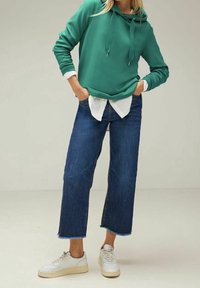 Zelená mikina s kapucí a šňůrkami přes bílou košili. Tmavě modré džíny s širokými nohavicemi a bílé tenisky s béžovými podrážkami.