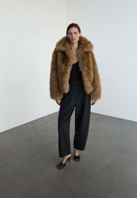 Hnedý kožený kabát z umelého kožúška so širokým golierom, skombinovaný s čiernymi nohavicami s vysokým pásom a čiernymi lodičkami s otvorenou špičkou, na jednoduchom pozadí.