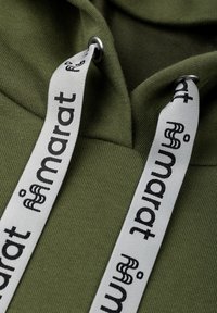 Lähikuva oliivinvihreästä kankaasta, jossa valkoiset kiristysnauhat, musta "namarat"-teksti ja metalliset silmukat.