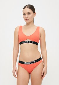 Gestreifter orangener Bikini aus zwei Teilen mit einem V-Ausschnitt-Top und passenden Bottoms. Schwarzes Calvin Klein Logo-Band um die Taille. Glatte Textur.