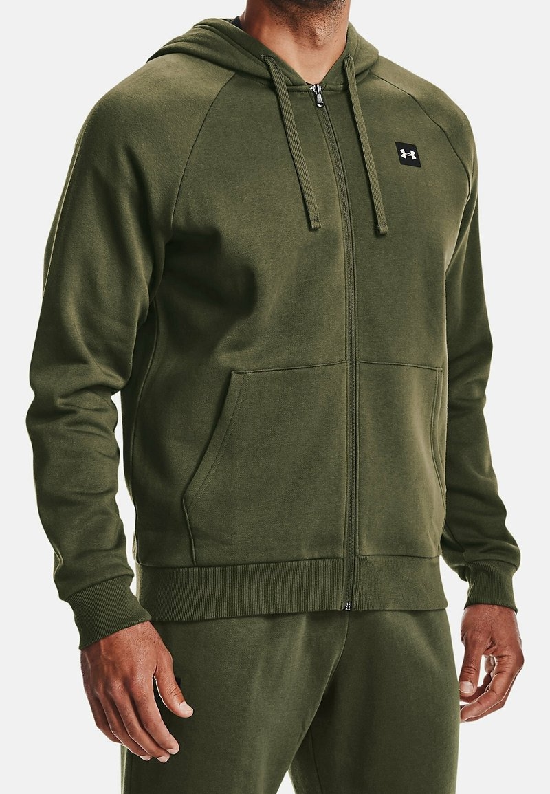 Homme portant un sweat à capuche zippé vert olive avec poches avant et cordons, montrant uniquement le haut du torse et les bras.