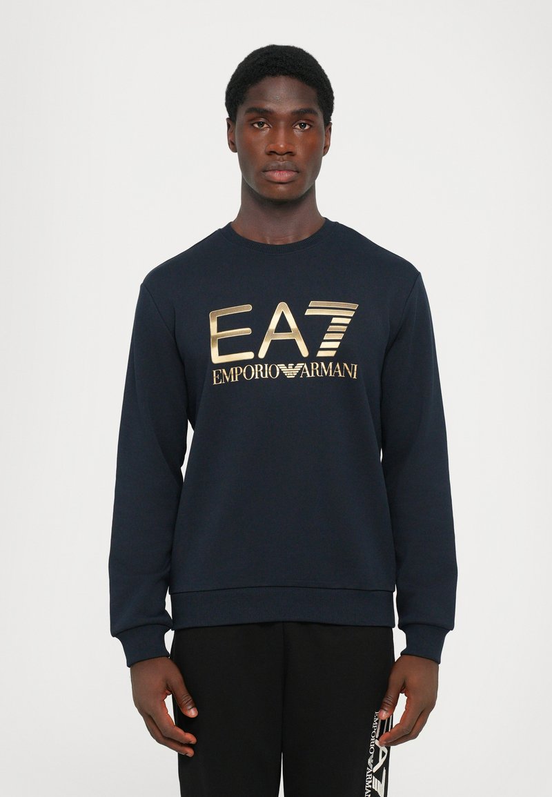 Navy sweatshirt with long sleeves and a crew neckline features a gold printed logo "EA7 EMPORIO ARMANI" on the front. Soft texture.