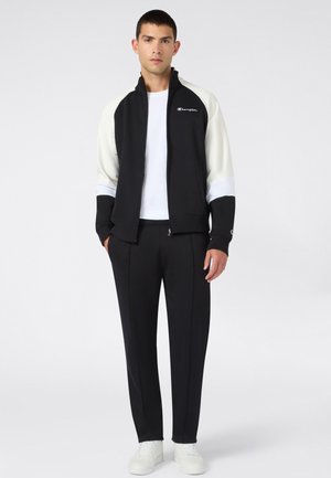Joven de pie con una chaqueta negra y blanca con cremallera sobre una camiseta blanca, pantalones negros y zapatillas blancas sobre un fondo liso y claro.