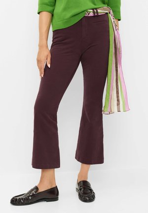 Kvinde iført mørkelilla cropped bukser, sorte loafers, grøn sweater og et stribet grønt, pink og brunt tørklæde bundet om livet.