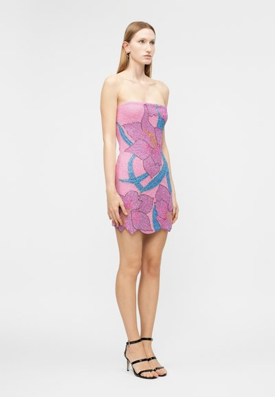 Ženska, ki stoji, oblečena v roza kratko obleko brez naramnic z velikimi vijoličnimi in modrimi cvetličnimi vzorci ter črnimi sandali na visoki peti z naramnicami.