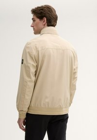 Beige lichtgewicht jack met een hoge kraag, ribgebreide mouwen en een soepele textuur. Bevat een logo-accent op de linker mouw.
