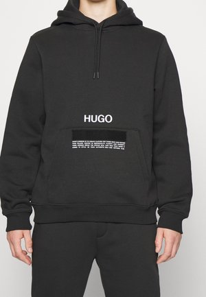 Czarna bluza z kapturem z przednią kieszenią typu kangur, biały napis "HUGO" nad mniejszym białym akapitem na kieszeni, noszona przez osobę od ramion do bioder.