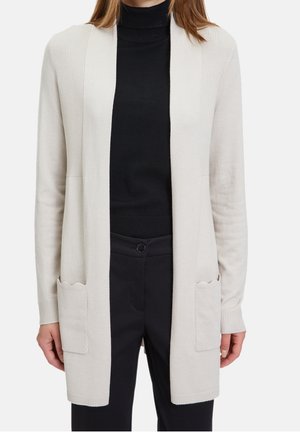 Femme portant un long cardigan beige avec des poches avant sur un col roulé noir et un pantalon noir, debout devant un fond blanc.