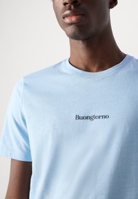 Hele sinine puuvillane T-särk, mis on ümarate kaelustega ja lühikeste varrukatega, millel on rinnal tumedates tähtedes trükitud "Buongiorno".
