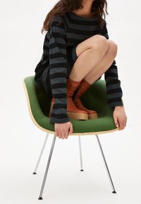 Zakřivená zelená židle se dřevěným rámem a kovovými nohami. Osoba v černobíle pruhovaném dlouhém svršku a hnědých vysokých botách po kolena.