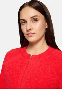 Czerwony, miękki sweter na zamek błyskawiczny z okrągłym dekoltem, wyposażony w metalowy zamek i gładką fakturę. Zbliżenie podkreśla jakość materiału.