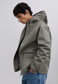 Chaqueta con capucha gris, con cremallera frontal, dos bolsillos delanteros y una textura suave. Combinada con jeans azules, vista de lado.