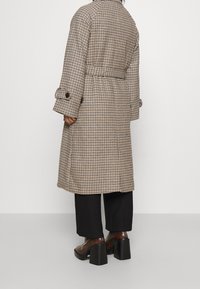 Lange, geruite jas in beige en bruin, met een riem in de taille en knoopdetails op de mouwen. Draagt over zwarte wijde broek en laarzen met hak.