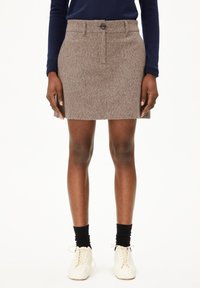 Femme portant une mini-jupe en laine marron, des cha socks noires, des chaussures blanches à lacets et un haut à manches longues bleu marine, se tenant devant un fond blanc.