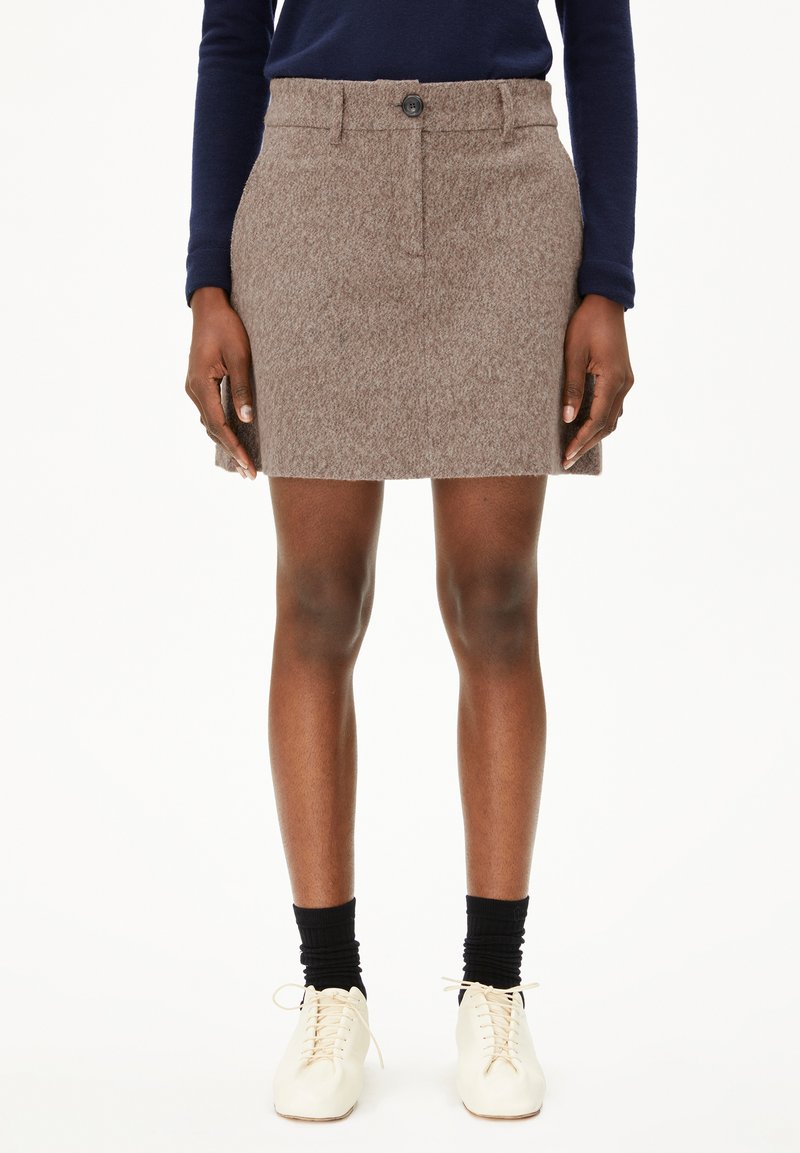 Femme portant une mini-jupe en laine marron, des cha socks noires, des chaussures blanches à lacets et un haut à manches longues bleu marine, se tenant devant un fond blanc.