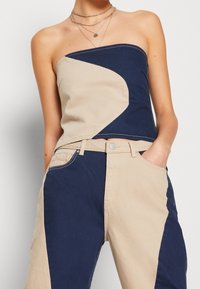 Top senza spalline con un design color block curvo in crema e blu navy. Tessuto attorcigliato nella parte superiore, indossato con pantaloni in denim beige dotati di tasche laterali.
