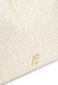 Μπεζ ύφασμα με υφή και επαναλαμβανόμενο γεωμετρικό μοτίβο, συνοδευόμενο από ένα μικρό χρυσό και λευκό διαπλεκόμενο λογότυπο "TH".