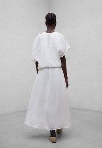 Femme aux cheveux tressés portant une robe blanche ample et des chaussures beiges, debout contre un fond gris clair uni, de dos.