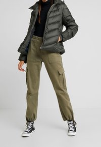 Doudoune vert olive foncé avec capuche et fermeture éclair, accompagnée d'un pantalon cargo beige avec poches latérales et chevilles resserrées. Baskets noires.