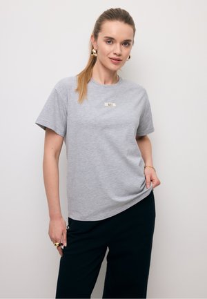 Młoda kobieta z długimi włosami związanymi w kucyk, ubrana w szary krótki rękaw i czarne spodnie, ze złotymi kolczykami kołami, pierścionkiem i bransoletką, stojąca na tle białej ściany.