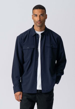 Marineblå skjorte med knapper lavet af ensfarvet stof, med to brystlommer og lange ærmer, lagdelt over en hvid t-shirt.