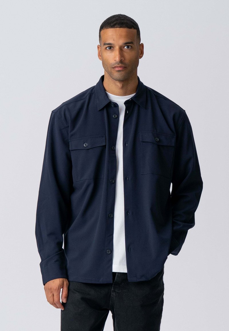 Marineblå skjorte med knapper lavet af ensfarvet stof, med to brystlommer og lange ærmer, lagdelt over en hvid t-shirt.