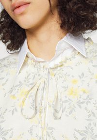 Blommönstrad topp med gula och grå blommor på en vit bakgrund. Har en volangkrage och knutdetaljer. Vit skjorta med krage under.