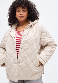 Bež prešita jakna z ruto, z zatiči na pritrditev, stranskimi žepi in sproščenim krojem, nošena preko rdeče-bele črtaste majice.