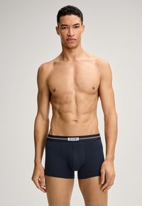 Modello maschile senza maglietta con fisico in forma, che indossa boxer blu navy con cintura nera e logo JOOP!, in piedi contro uno sfondo neutro.