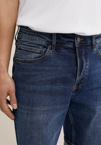 Tamsiai mėlynos džinsinės kelnės su liesu siluetu, turinčios vidutinį liemenį, priekinius kišenes ir kontrastingą siūlę. Lygi tekstūra su lengvu išblukimu.