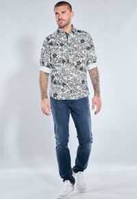 Schwarz-weiß gemustertes Hemd mit Blumenmuster, Ärmel hochgekrempelt, kombiniert mit dunkelblauen Jeans und hellen Sportschuhen.