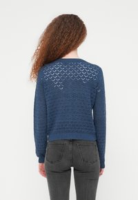 Πλεκτό πουλόβερ σε ναυτικό μπλε με δαντελωτό μοτίβο, ριμπ μανσέτες και τελείωμα, παρουσιάζοντας υφασμένο σχέδιο. Φοριέται με σκούρο γκρι τζιν.